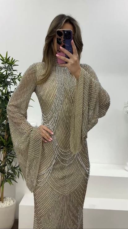 فستان سهرة كريمي اللون، بأكمام واسعة مزينة بتفاصيل دقيقة، وقصة متدلية مطرزة بالخرز 